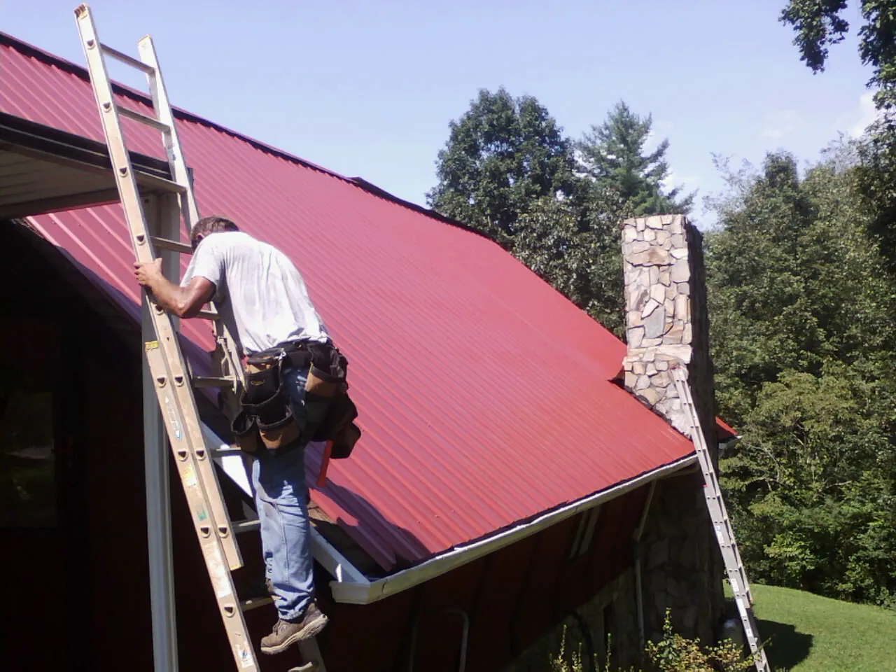 Roofer on a ladder beside a red metal roof and stone chimney — Soffit & Fascia Repair by Roofing Brothers in Hawaiian Gardens
